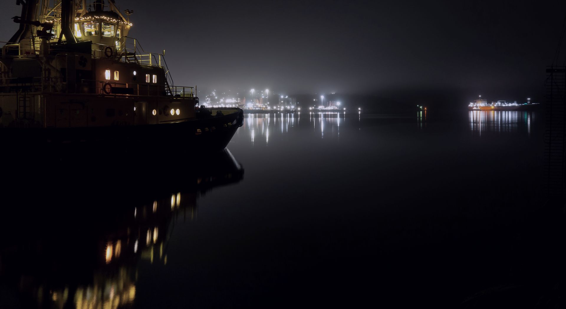 A boat moored on still black water at night, harbour lights glowing through the fog