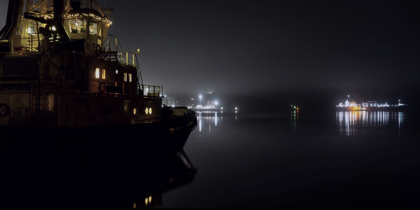 A boat's lit wheelhouse in fog at night, lights strung across the far shore